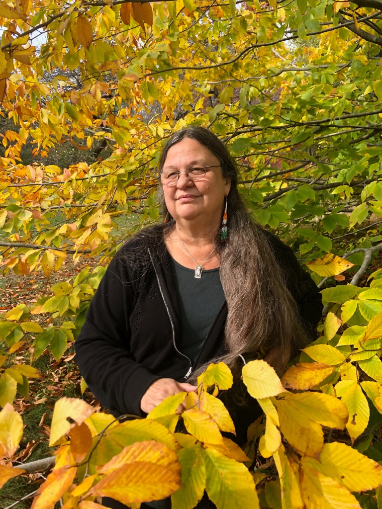 Judy Dow stands surrounded by yellow leaves, looking past the camera to the left with a slight smile.